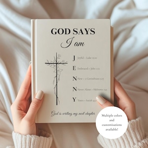 Puede incluir: Un libro de tapa dura color crema con el texto negro GOD SAYS I am, una ilustración de una cruz floral y el nombre JENNY con versículos bíblicos. El texto inferior dice God is writing my next chapter.