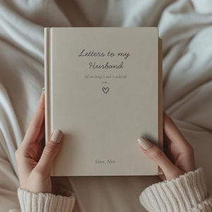 May include: A cream-coloured book titled "Letters to my Husband" with cursive text. The cover includes the text "All the things I wish I could tell you..." and a heart symbol. The book is held open by a person.