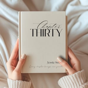 May include: A cream-coloured hardback book with black text on the front cover. The text reads Chapter THIRTY, Jessie Sm, and Every chapter brings new growth. The book has a smooth, matte finish and is held upright with both hands.