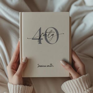 May include: A cream-coloured journal featuring the word "Forty" in grey script and the number "40" in a bold grey font. The name "Jessica Smith" is written below in a delicate script. The journal is held by a person.