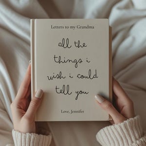 May include: A cream-coloured book titled "Letters to my Grandma" held by a person. The book's cover features the handwritten text "all the things i wish i could tell you" and "Love, Jennifer". The book has a simple, elegant design.