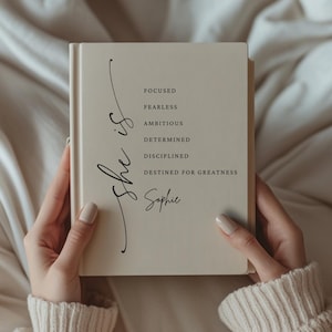 May include: A cream-colored journal with the words "She is" in cursive script. The journal lists the words "Focused, Fearless, Ambitious, Determined, Disciplined, Destined for Greatness" and the name "Sophie".