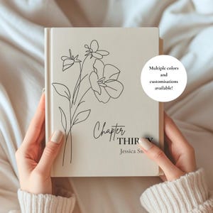 May include: A cream-colored hardcover book featuring a black line-art floral illustration on the front cover. The text reads Chapter THIRTY and Jessica Sn. A circular white graphic states Multiple colors and customisations available!