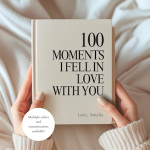 May include: A cream-coloured hardcover book with black text that reads 100 MOMENTS I FELL IN LOVE WITH YOU and Love, Amelia. A circular white graphic in the bottom left corner contains the text Multiple colours and customisations available!