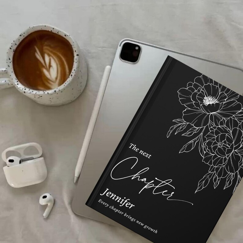 May include: A black journal with white floral illustrations and the text "The next Chapter Jennifer". A cup of coffee with latte art, a tablet, a stylus, and wireless earbuds are also in the image.