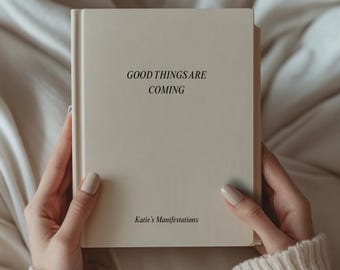 May include: A cream-colored book with the text "GOOD THINGS ARE COMING" in black. The book is held by a person wearing a cream-colored sweater. The text "Katie's Manifestations" is printed at the bottom of the book.
