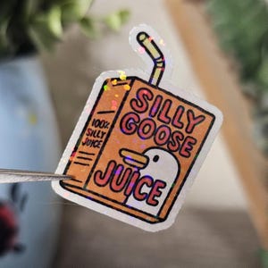 May include: A holographic sticker featuring a juice box design with the words "SILLY GOOSE JUICE" in orange and white. The juice box has a cartoon goose illustration and a straw.