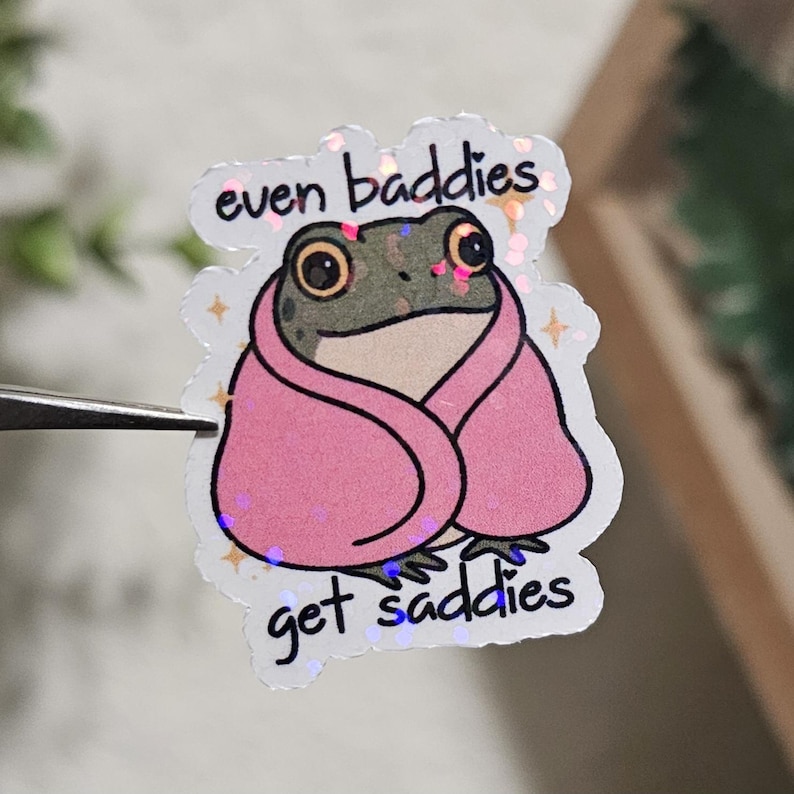 May include: A sticker featuring a cartoon frog wrapped in a pink blanket. The frog has large eyes and is green and gray. The sticker has the text "even baddies get saddies" in black lettering.