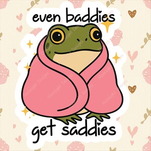 May include: A sticker featuring a cartoon frog wrapped in a pink blanket. The frog has large yellow eyes and is set against a cream background with hearts and stars. Text reads "even baddies get saddies."