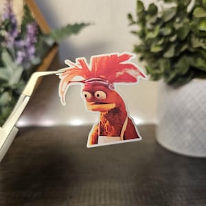 Może przedstawiać: Naklejka przedstawiająca czerwonowłosą marionetkę z zaskoczonym wyrazem twarzy. Marionetka ma na sobie biały fartuch i ma czerwono-pomarańczowe ciało.
