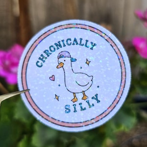 May include: A circular sticker with a holographic finish featuring a white duck wearing a blue baseball cap. The text "CHRONICALLY SILLY" is printed in blue around the duck, accompanied by small blue stars and a pink heart on a white background.