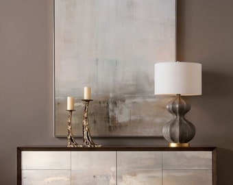 Puede incluir: Un aparador de madera oscura con cuatro cajones y cuatro puertas. Las puertas y los cajones tienen un acabado pintado de gris claro con un diseño abstracto sutil. El aparador está decorado con dos candelabros dorados y una lámpara de mesa de cerámica gris con una pantalla blanca.