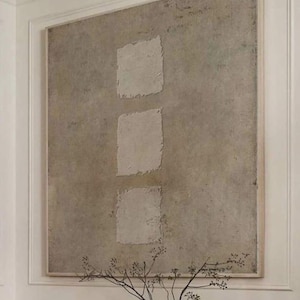 Może przedstawiać: Duży abstrakcyjny obraz z trzema teksturowanymi, kwadratowymi kształtami w neutralnej palecie kolorów. Dzieło sztuki jest oprawione i zamontowane na białej ścianie. Poniżej znajduje się dekoracyjny wazon z gałęziami.