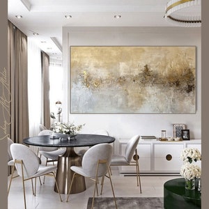 Op de afbeelding: Een moderne eetkamer met een ronde zwarte marmeren tafel en een gouden metalen onderstel. De kamer heeft een groot abstract schilderij met gouden en bruine accenten, een witte kast met gouden accenten en een ronde groene bijzettafel met witte bloemen.