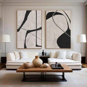 以下が含まれることがあります： テクスチャ加工された白い背景に黒い線と形が描かれた2つの大きな抽象画が、白いソファの上に飾られています。木製のコーヒーテーブルには装飾的な花瓶と黒いトレイがあり、ソファの前に置かれています。2つのフロアランプがソファの両側にあります。