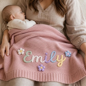 May include: A dusty rose knitted baby blanket featuring the name Emily embroidered in pastel-coloured yarn. The blanket is decorated with small, three-dimensional flower appliques in white, blue, and purple, and has a ribbed border along the bottom edge.