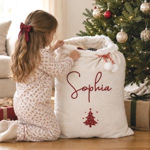 Pode incluir: Um saco de presente de Natal grande em tecido branco com uma borda de pele sintética branca no topo, cordões vermelhos com pompons e o nome Sophia bordado em vermelho sobre um pequeno gráfico vermelho de uma árvore de Natal decorada.