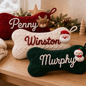Op de afbeelding: Drie botvormige gebreide kerstsokken in donkerrood, crème en donkergroen. Elke sok heeft een ophanglus en een gepersonaliseerde naam in cursief schrift: Penny met een rendier, Winston met een kerstman en Murphy met een sneeuwpopapplicatie.