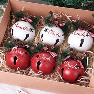 Puede incluir: Una caja de adornos de campanas navideñas personalizadas. Tres campanas blancas con los nombres Amelia, Olivia y Emily, y tres campanas rojas con Harry, Jack y Charlie. Cada campana está decorada con pino y un lazo a cuadros rojo y blanco.
