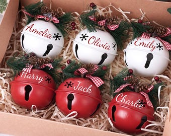 May include: A box of personalized Christmas bell ornaments. Three white bells with names Amelia, Olivia, and Emily, and three red bells with names Harry, Jack, and Charlie. Each bell is decorated with pine and a red and white checkered bow.