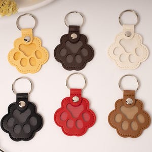 May include: Six paw-shaped keychains in various colors: yellow, brown, off-white, black, red, and tan. Each keychain features a metal ring and a matching leatherette paw print design, perfect for pet lovers.