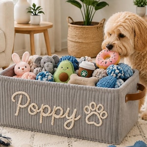 Peut inclure: Un bac de rangement rectangulaire en velours côtelé gris avec des poignées en cuir marron, portant le nom Poppy et une empreinte de patte brodés en corde blanche. Le bac est rempli de divers jouets en peluche, dont un avocat et un donut.