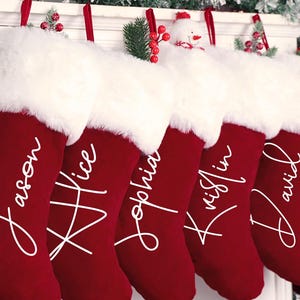 May include: Six red Christmas stockings with white faux fur cuffs hang from a white mantel. Each stocking is personalized with a name in white script: Jason, Alice, Sophia, Kristin, and David. Festive holiday decor surrounds the mantel.