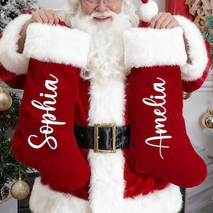 Puede incluir: Papá Noel sosteniendo dos calcetines navideños rojos con ribete de piel sintética blanca. Los calcetines están personalizados con los nombres "Sophia" y "Amelia" en escritura blanca. La escena es festiva con árboles de Navidad y decoraciones.
