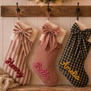 May include: Three hanging Christmas stockings with personalised names embroidered in yarn. The first is red and white striped with a red bow and the name Henry. The second is solid dusty pink with a matching bow and the name Bella. The third is black and white gingham with a matching bow and the name Amelia.