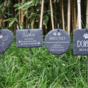 May include: Four dark grey slate pet memorial markers in various shapes, each with a metal stake. The markers are engraved with pet names, dates, and memorial phrases. The markers are in a grassy area.