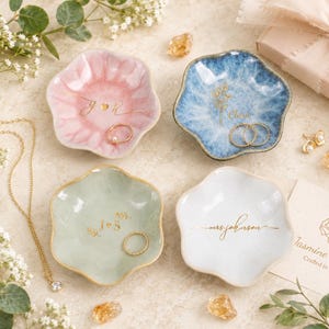 May include: Four scalloped ceramic trinket dishes in pastel pink, blue, sage green, and white. Each dish features gold-colored custom lettering or floral designs. The dishes are arranged with gold jewelry, small crystals, and a branded card.