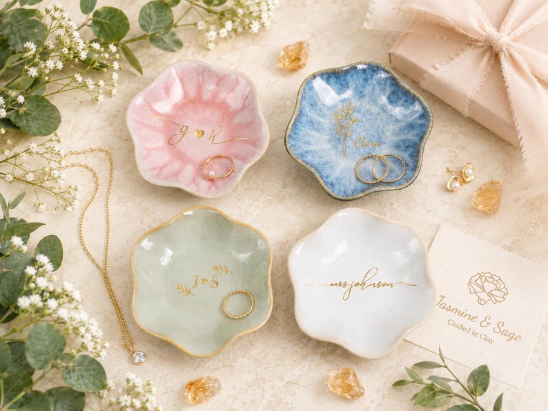 May include: Four scalloped ceramic trinket dishes in pastel pink, blue, sage green, and white. Each dish features gold-colored custom lettering or floral designs. The dishes are arranged with gold jewelry, small crystals, and a branded card.