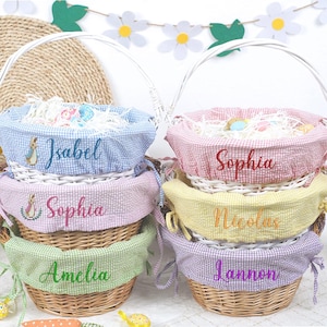 May include: Wicker Easter baskets with white handles and colourful gingham liners. Each basket has a name embroidered on the liner: Isabel, Sophia, Amelia, Nicolas, and Lannon. The baskets are filled with decorative Easter grass.