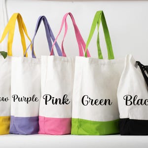 May include: Five canvas tote bags with coloured bottoms and handles. The bags are white with yellow, purple, pink, green, and black accents. Each bag has the colour name printed on the front in a cursive font. The bags are standing upright.