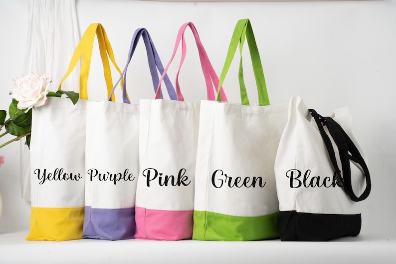 May include: Five canvas tote bags with coloured bottoms and handles. The bags are white with yellow, purple, pink, green, and black accents. Each bag has the colour name printed on the front in a cursive font. The bags are standing upright.