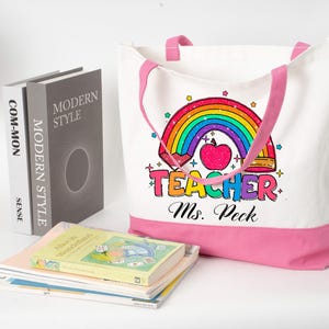 May include: White canvas tote bag with pink trim and handles, featuring a rainbow and apple design with the word "Teacher" and the name "Ms. Peck". Books are stacked nearby. The bag is a practical accessory.