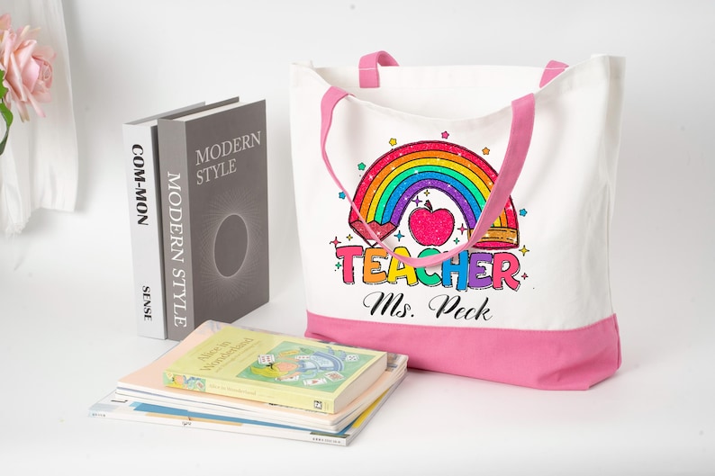 May include: White canvas tote bag with pink trim and handles, featuring a rainbow and apple design with the word "Teacher" and the name "Ms. Peck". Books are stacked nearby. The bag is a practical accessory.