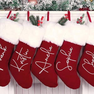 Könnte beinhalten: Fünf rote Weihnachtsstrümpfe mit weißem Fellbesatz und personalisierten Namen in weißer Schrift. Jeder Strumpf ist mit einem Zweig aus Grün und roten Beeren verziert, der an einem weißen Kaminsims hängt.