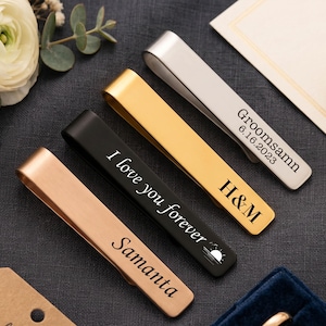 Puede incluir: Cuatro pasadores de corbata de metal en acabados oro rosa, negro, oro y plata. El pasador de oro rosa está grabado con Samanta, el negro con I love you forever y un icono de sol, el dorado con H&M y el plateado con Groomsamn 6.16.2023.