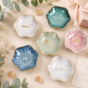 May include: Six small, flower-shaped ceramic trinket dishes in various colors including white, teal, sage green, pink, and blue. Each dish features gold or white personalized text or monograms. A small white card with the text thank you is included.