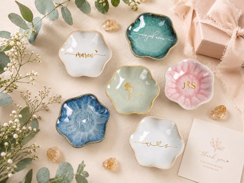 May include: Six small, flower-shaped ceramic trinket dishes in various colors including white, teal, sage green, pink, and blue. Each dish features gold or white personalized text or monograms. A small white card with the text thank you is included.