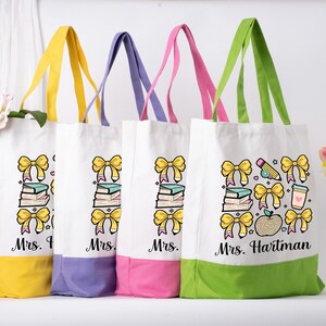 May include: Four white canvas tote bags with coloured handles and bases. Each bag features a design of yellow bows, books, and the name "Mrs." followed by a name. One bag has a pencil, coffee cup, and apple.
