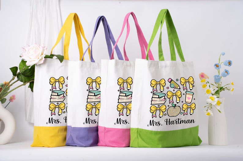 May include: Four white canvas tote bags with coloured handles and bases. Each bag features a design of yellow bows, books, and the name "Mrs." followed by a name. One bag has a pencil, coffee cup, and apple.