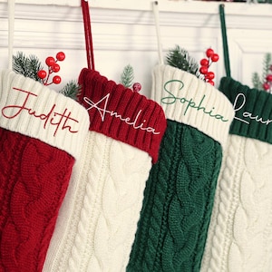 May include: Four cable-knit Christmas stockings in red, white, and green. Each stocking features a white cuff with a name embroidered in a matching colour: Judith, Amelia, Sophia, and Lauren. Greenery sprigs and red berries decorate the tops.