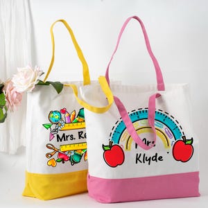 May include: Two white canvas tote bags with colourful designs and handles. One bag has a yellow base and handle, the other has a pink base and handle. The bags feature school-themed graphics, including apples, rainbows, and rulers, with the text "Mrs. R" and "Mrs. Klyde".