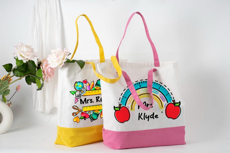 May include: Two white canvas tote bags with colourful designs and handles. One bag has a yellow base and handle, the other has a pink base and handle. The bags feature school-themed graphics, including apples, rainbows, and rulers, with the text "Mrs. R" and "Mrs. Klyde".