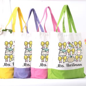 Op de afbeelding: Vier witte tote bags met kleurrijke accenten. Elke tas heeft een handvat en een onderste paneel in een andere kleur: geel, paars, roze en groen. De tassen hebben een thema-ontwerp voor leraren met strikken, boeken en de naam "Mrs. Hartman".