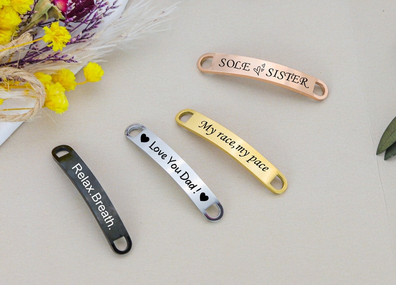 May include: Four shoe charms with different engraved messages. The charms are made of metal and have a silver, gold, and rose gold finish. The messages are "Sole Sister", "My race, my pace", "Love You Dad!" and "Relax. Breath."