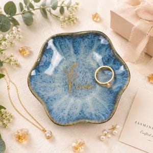 May include: A scalloped, blue and white glazed ceramic trinket dish featuring a gold-colored botanical illustration and the name Evelyn printed in gold. A gold ring rests inside the dish, which is surrounded by a gold necklace and small pearls.