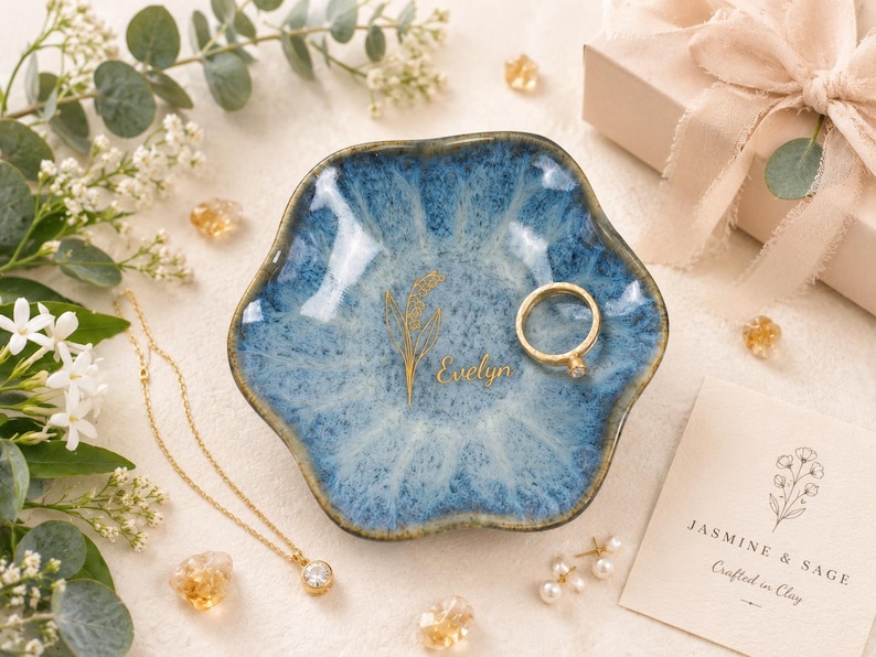 May include: A scalloped, blue and white glazed ceramic trinket dish featuring a gold-colored botanical illustration and the name Evelyn printed in gold. A gold ring rests inside the dish, which is surrounded by a gold necklace and small pearls.